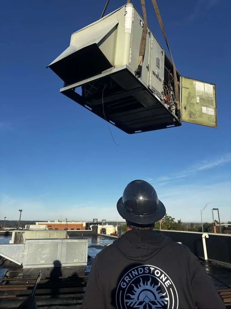 Technician in a Grindstone hoodie on a roof, guiding a crane cable as a rooftop HVAC unit is lifted into place.
