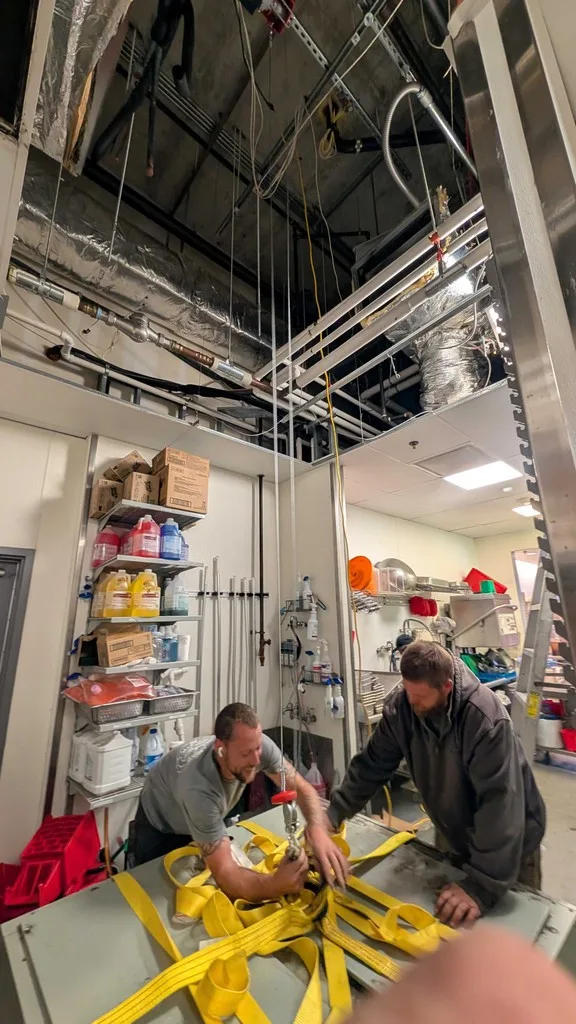 Two technicians on ladders rigging equipment through a commercial kitchen ceiling.