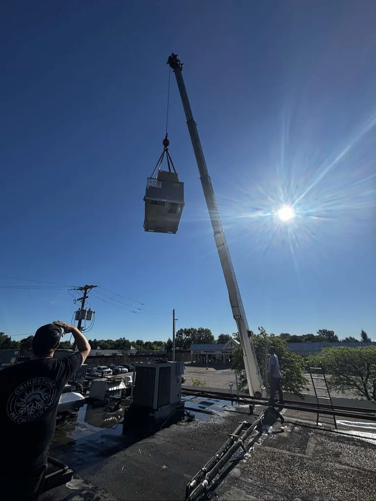 Crane lifting a rooftop unit into the sun, with the Grindstone logo on a worker's hoodie.