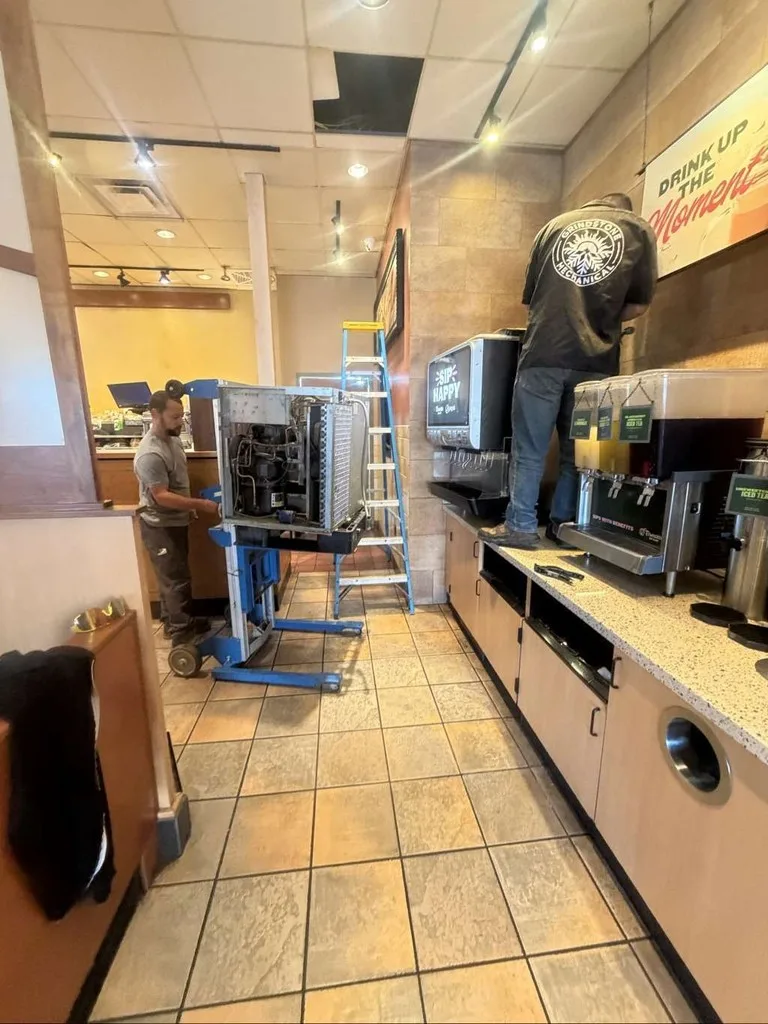 Technician on a ladder servicing refrigeration equipment behind a restaurant beverage counter.
