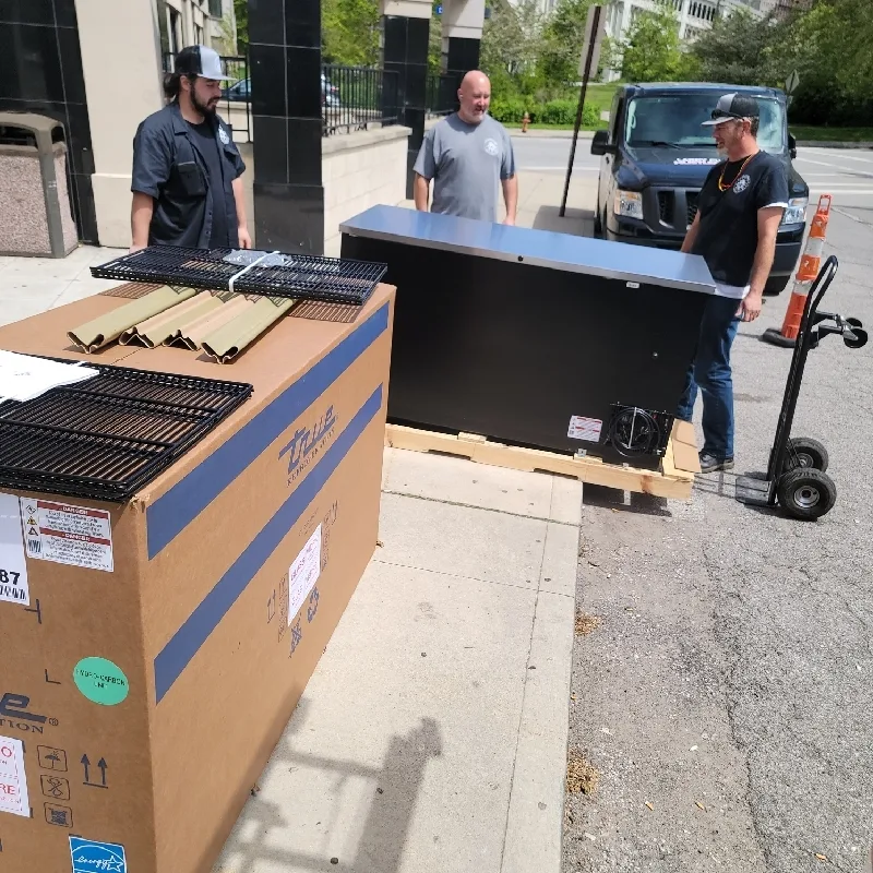 Two crew members unboxing a new stainless True refrigerator at the curb beside the Grindstone van.