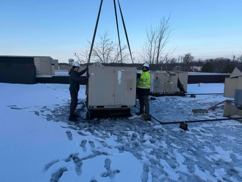 Two hard-hatted technicians guiding a rooftop unit into place on a snowy roof.