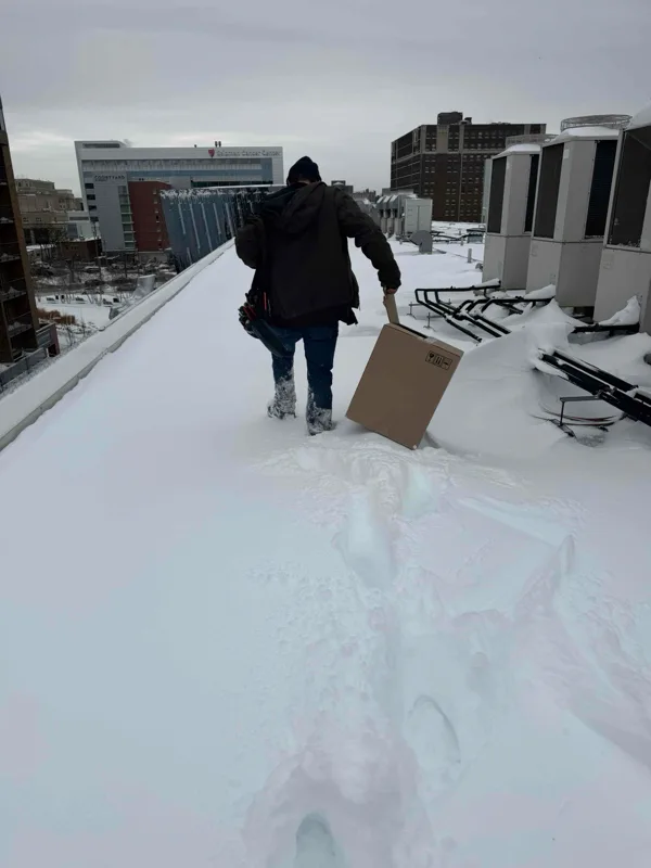 Technician walking through snow on a rooftop toward a pile of cardboard parts boxes.